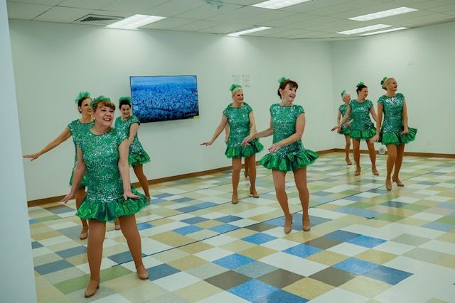 A group of dancers in matching green dresses perform in a practice room with a checkered floor and a wall-mounted TV.