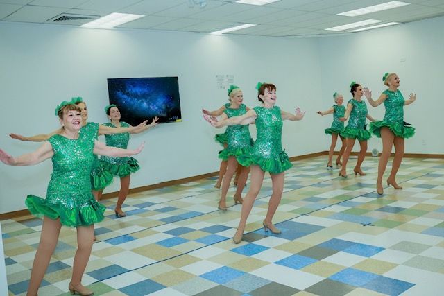 A group of performers in matching green sequined dresses dancing in a bright room with a checkered floor.