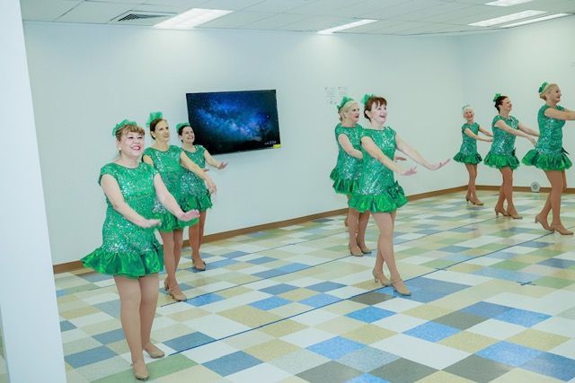 A group of people wearing green sequined dance costumes perform a routine in a bright, tiled-floor studio.