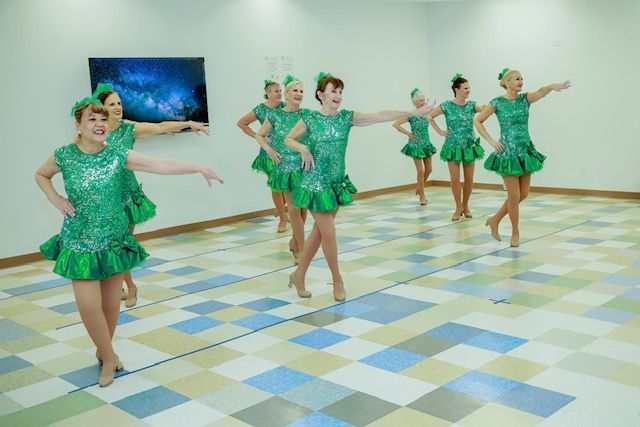 A group of dancers in matching green sequined dresses performs a dance routine in a studio with a patterned tile floor.