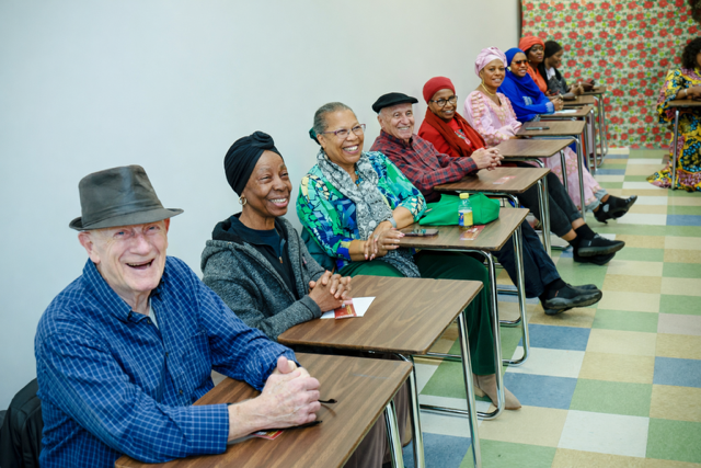 A group of people sit at desks in a classroom, smiling and facing forward, arranged in a row in a bright, tiled room.
