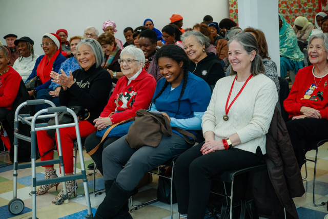 A group of people sitting indoors at an event, clapping and watching a presentation, with a walker in the foreground.