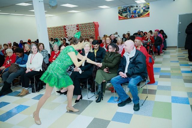 A performer in a bright green sequined dress dances in front of an audience seated in a brightly lit community room.