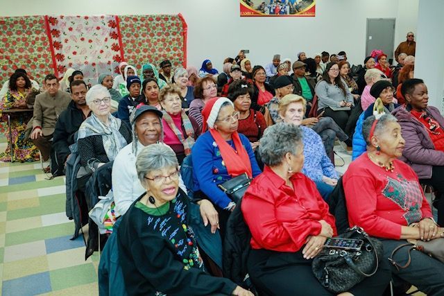 A group of people sitting in chairs in a hall for an event, many wearing red and holiday-themed attire.