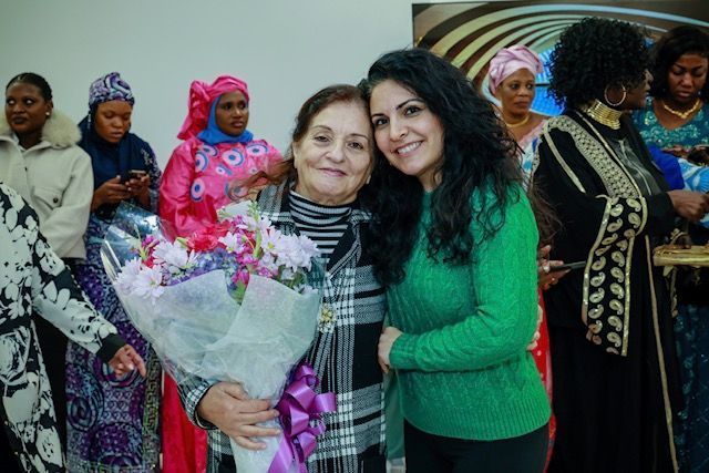 Two women smiling closely together, one holding a large bouquet of flowers, surrounded by a group in a room.