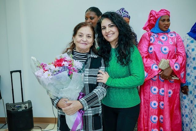 A woman in a green sweater smiles as she stands beside a woman in a plaid coat holding a large bouquet of flowers.