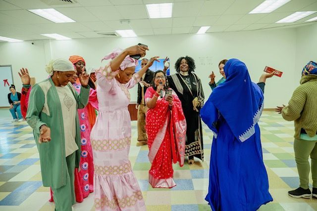People in diverse, colorful clothing dance together in a brightly lit indoor space with a checkered floor.
