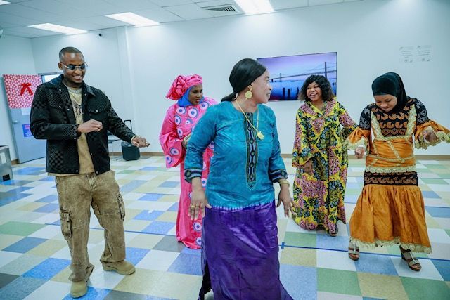 Five people in diverse, colorful traditional attire dance together in a brightly lit, tiled-floor multipurpose room.