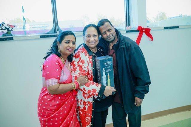 Three people stand indoors, one holding a gift box. Two wear formal attire, one wears a jacket. A red bow is on the wall.