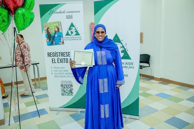 A smiling person in a blue hijab and matching dress holds a certificate in front of two branded green and white banners.