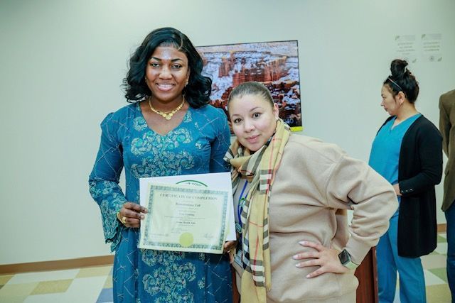 Two people pose together, one holding a certificate, in an indoor setting with a person visible in the background.