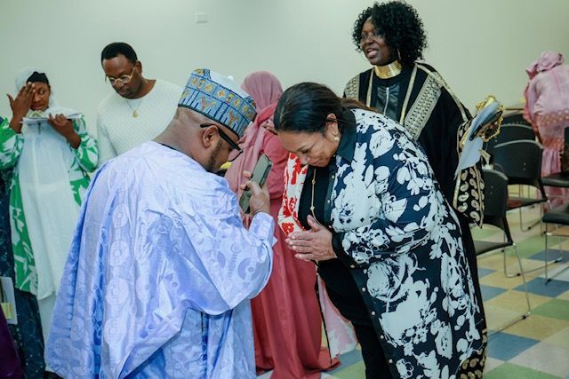 A man in a light blue traditional robe and cap bows toward a woman in a black and white floral jacket, greeting her.