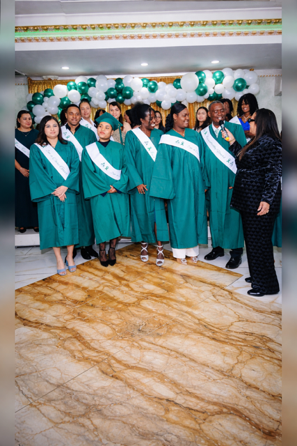 Graduates in green gowns and white sashes stand on a patterned floor while a person speaks to them in a decorated room.