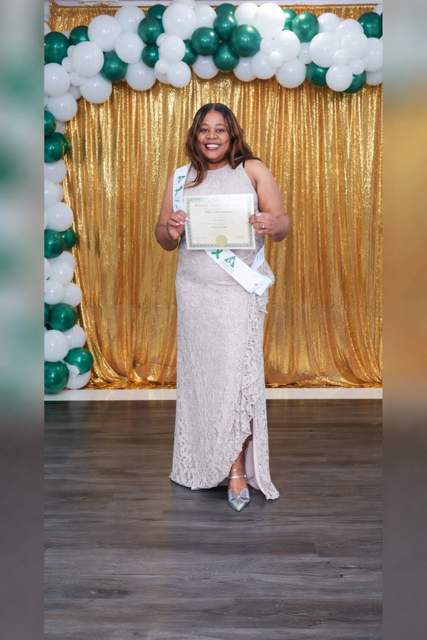 A person in a floor-length gown stands smiling before a gold curtain and balloon arch, holding a certificate.