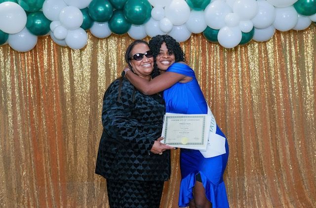 Two people embrace in front of a gold backdrop and green-and-white balloons; one holds a certificate.