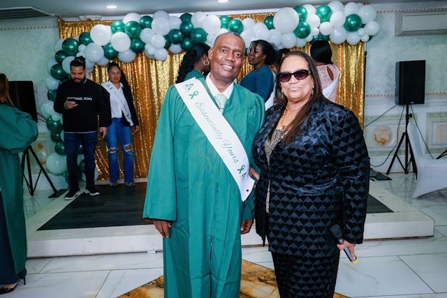 A graduate wearing a green gown and sash stands smiling next to a woman in a black patterned blazer at an indoor event.