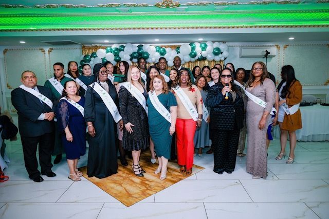 A diverse group in formal attire, some wearing sashes, poses for a photo in a decorated event hall with white balloons.