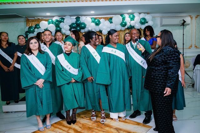 A group of people wearing green graduation robes and stoles stand indoors in front of a balloon arch as one speaks.