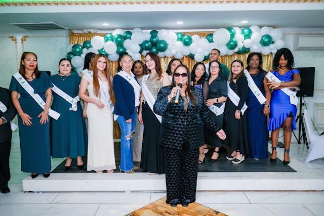 A woman speaks into a microphone before a line of participants in gowns and sashes at a formal indoor stage event.