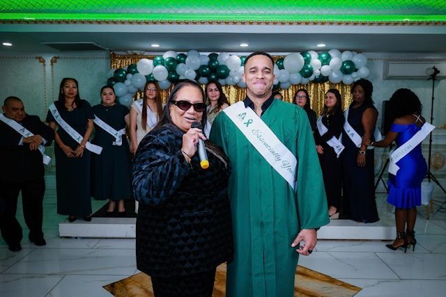 A person in a green robe and a woman with a microphone stand before a group in formal wear at a decorated event hall.