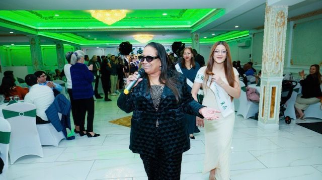 A speaker in a patterned black suit holds a microphone while a sash-wearing participant walks by in a bright event hall.