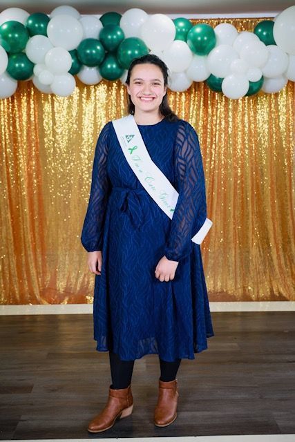 A smiling person in a blue long-sleeved dress and brown boots with a white sash stands before a gold curtain and balloons.