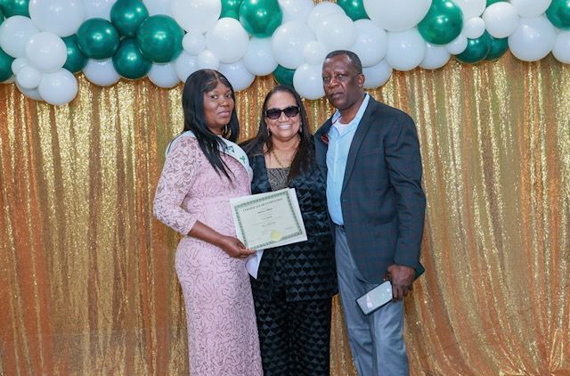 Three people stand before a gold curtain and green and white balloon arch; the center person holds a certificate.