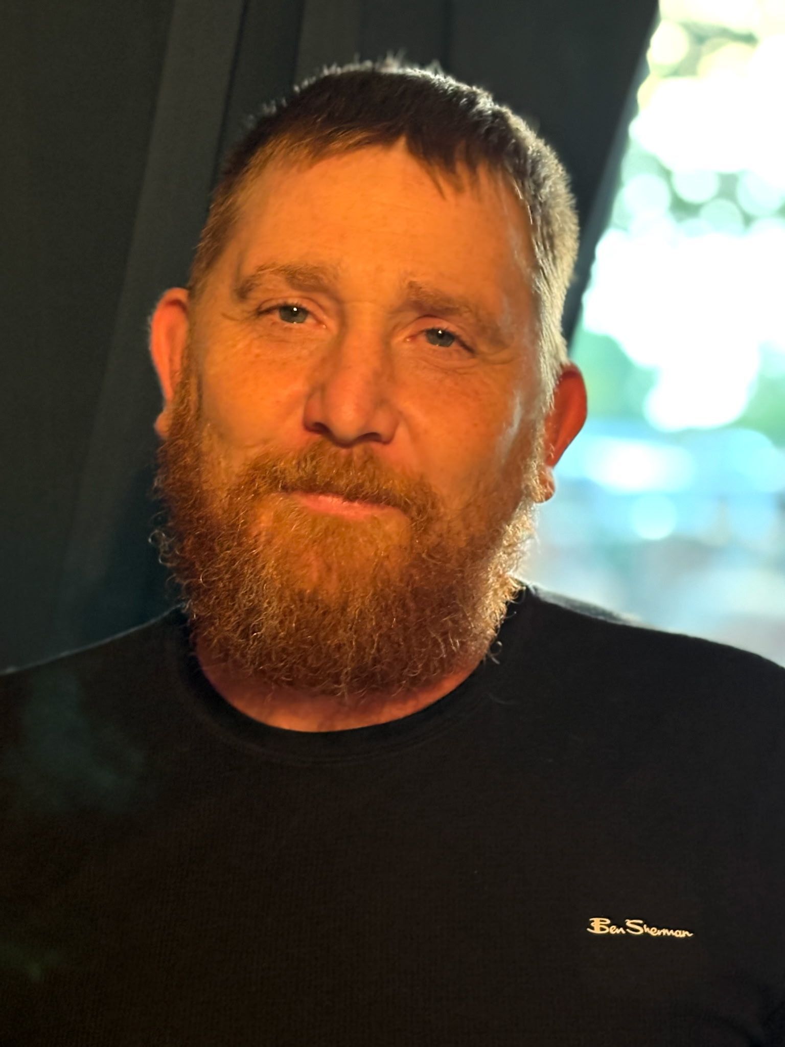 A person with a full beard and short hair wearing a black shirt, looking directly at the camera in a dimly lit setting.