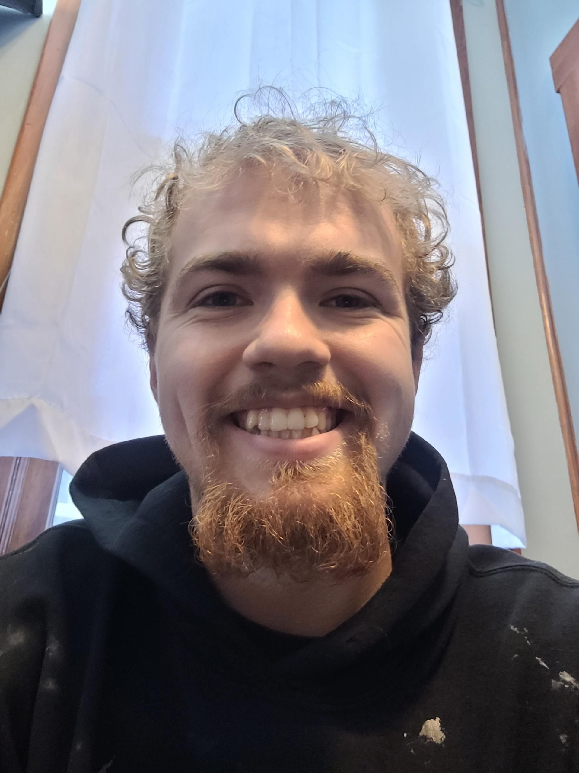 A person with curly blonde hair and a beard smiling at the camera, wearing a black hooded sweatshirt.