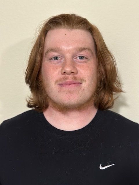 A person with shoulder-length reddish-brown hair, wearing a black crew-neck shirt, faces the camera against a beige wall.