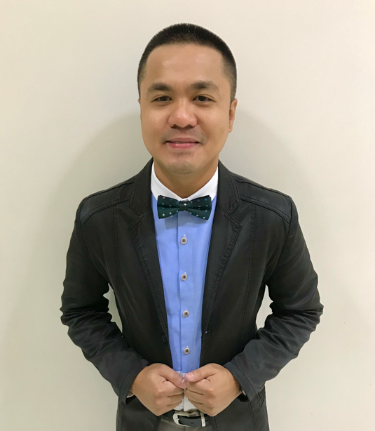 A person with a short haircut, wearing a blue shirt, patterned bowtie, and dark blazer, posing against a plain wall.