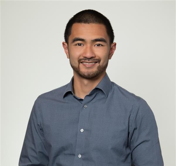 A man with short hair and a beard, wearing a blue button-down shirt, smiles against a plain light gray background.
