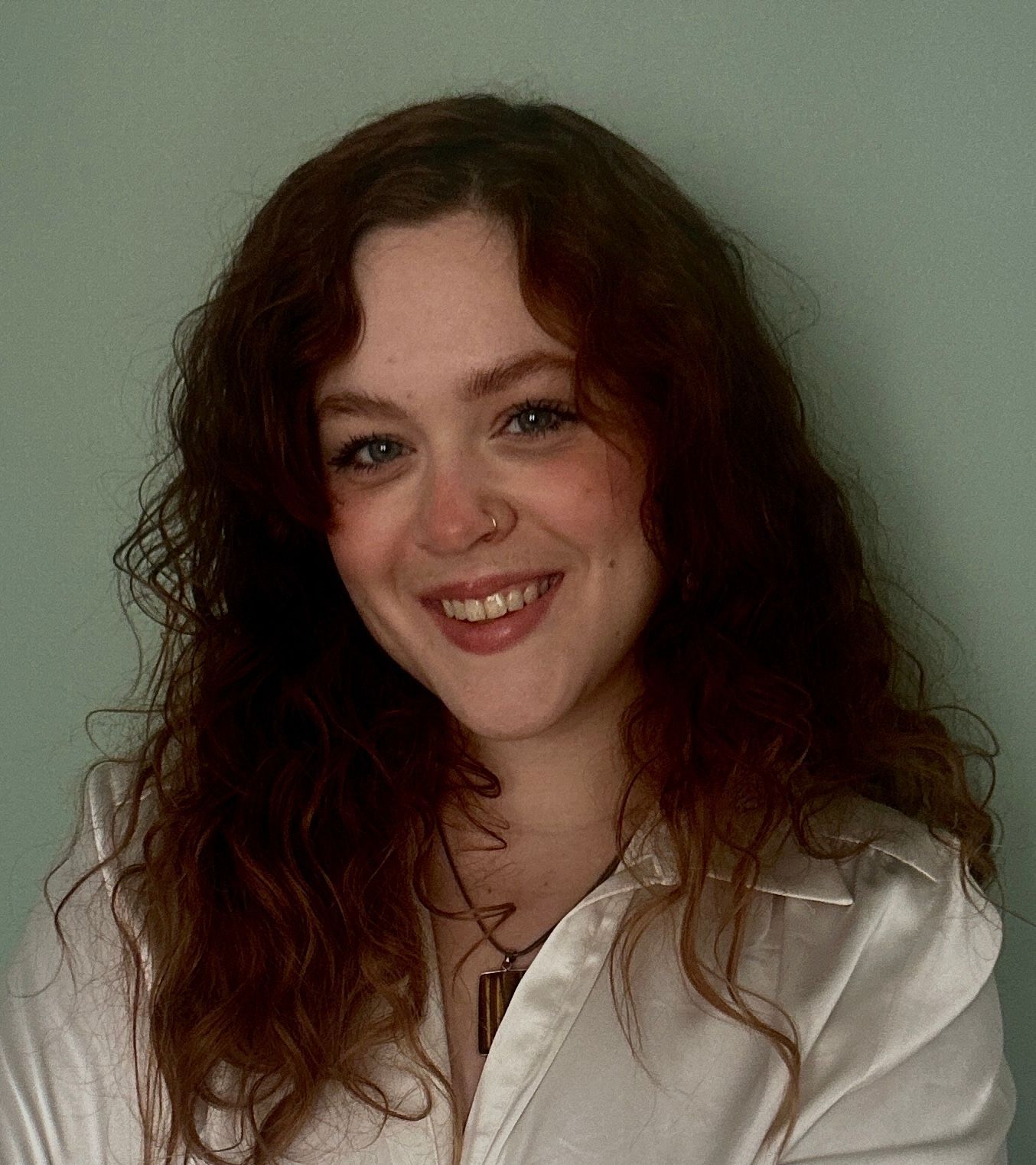 A smiling person with long, wavy auburn hair wearing a white collared shirt and a pendant necklace against a green wall.