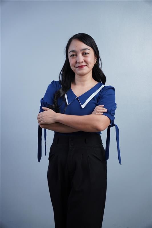 A person with long dark hair, wearing a blue collared blouse and black trousers, stands with arms crossed against a wall.