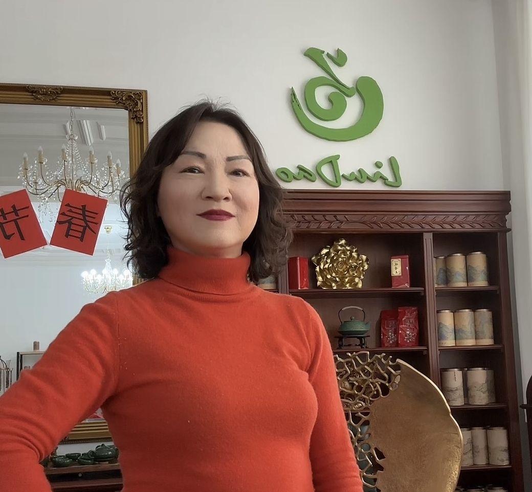 Eine Frau in einem orangefarbenen Rollkragenpullover steht vor einer Wand mit einem grünen Buchstaben G darauf