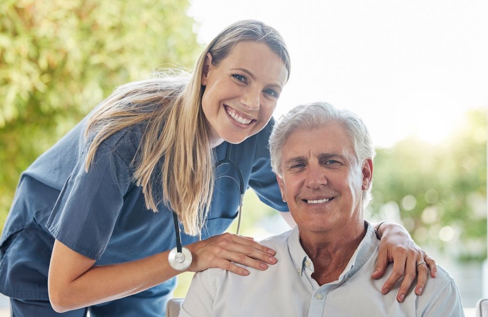 Woman in scrubs with stethoscope, smiling, arm around smiling older man. Outdoors with sunlight.