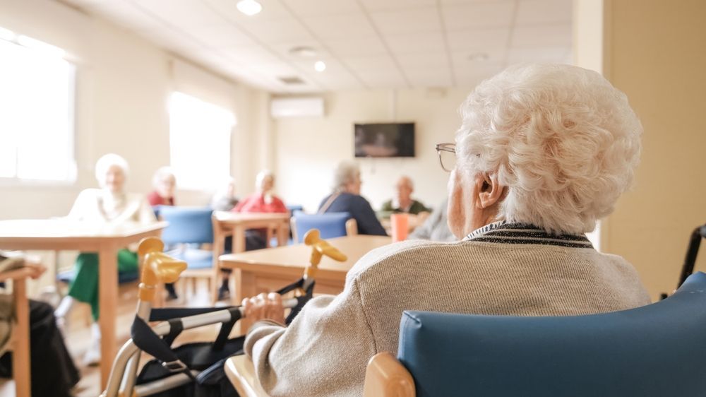 An elderly woman in a wheelchair, looking towards a group of people at tables in a brightly lit room.