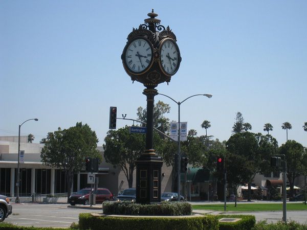 An ornate, vintage-style street clock stands on a pedestal in an outdoor plaza with trees and traffic lights in San Marino CA