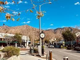 A street scene in Sierra Madre featuring a prominent clock on a pole, storefronts, and mountains under a blue sky.