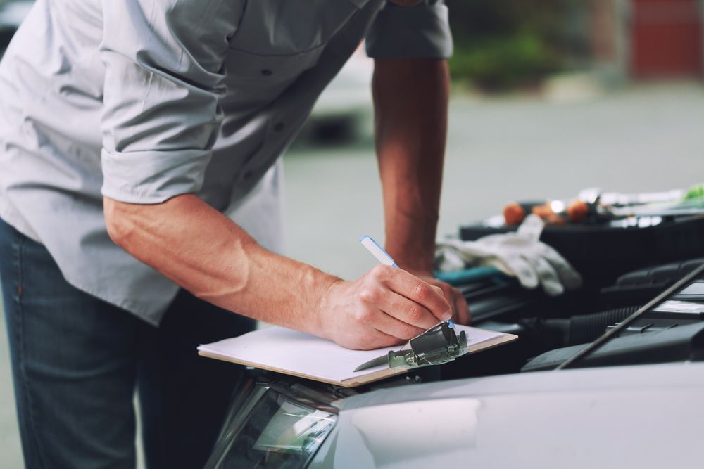 A Man Is Writing On A Clipboard While Looking Under The Hood Of A Car — CS Mechanical Repairs & Services In Wauchope, NSW