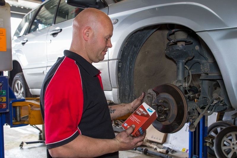 A Man Is Working On A Car In A Garage While Holding A Box Of Brake Pads — CS Mechanical Repairs & Services In Wauchope, NSW