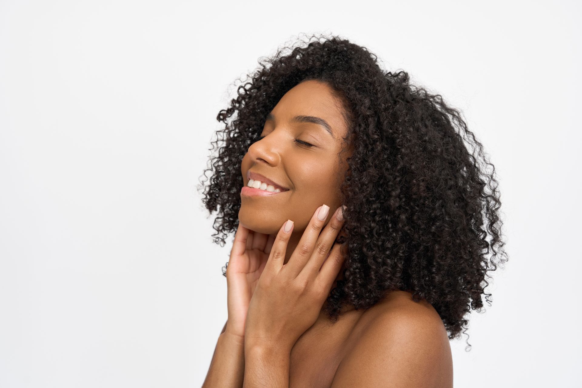 Smiling Black woman with curly hair, eyes closed, touching her neck against a white background.