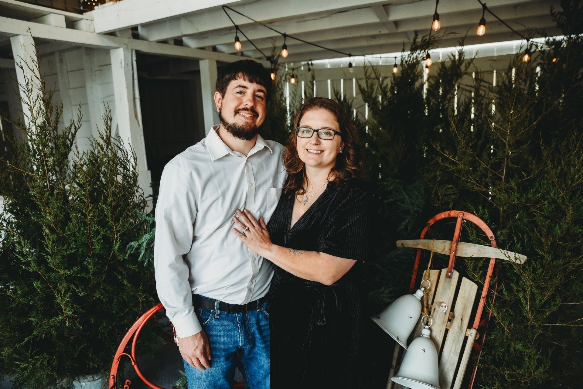 Couple standing together outdoors, smiling, with greenery and vintage sled.
