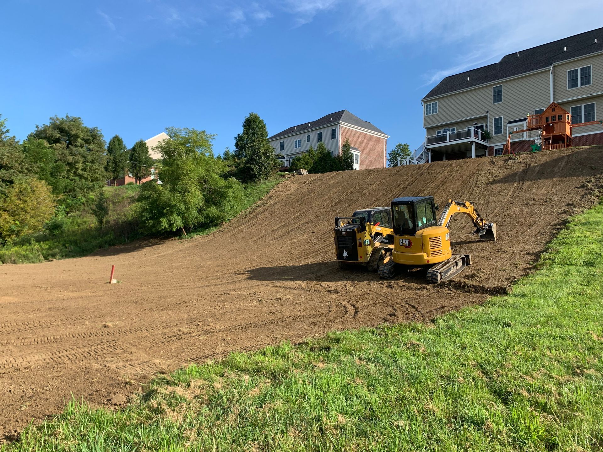 A yellow excavator is working on a dirt hill in a residential area.