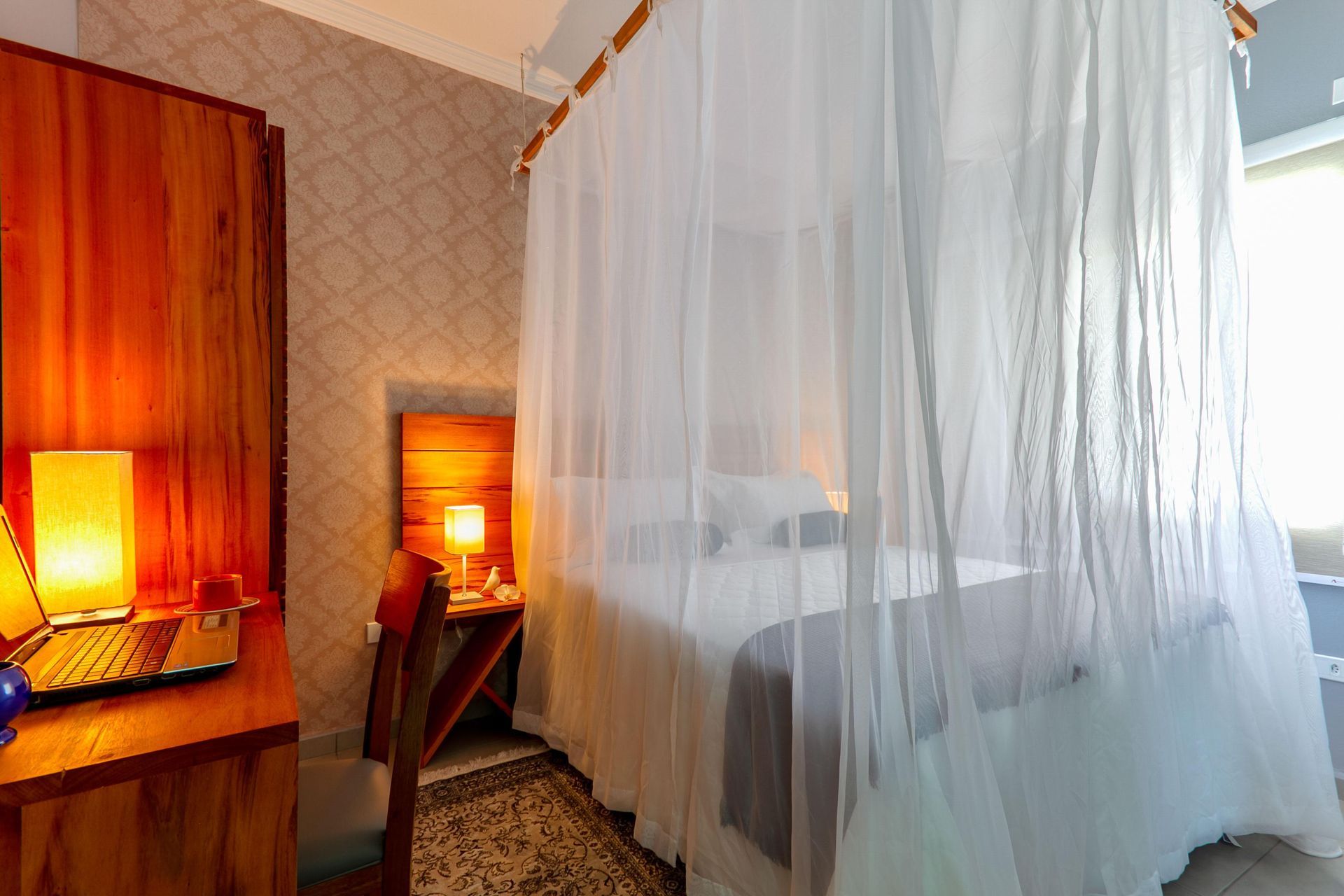 Quarto aconchegante com dossel branco translúcido, abajur de cabeceira com luz quente e móveis de madeira.