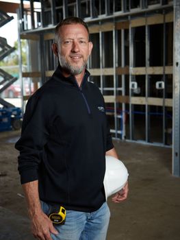 A man in a black sweater is holding a hard hat and a tape measure.