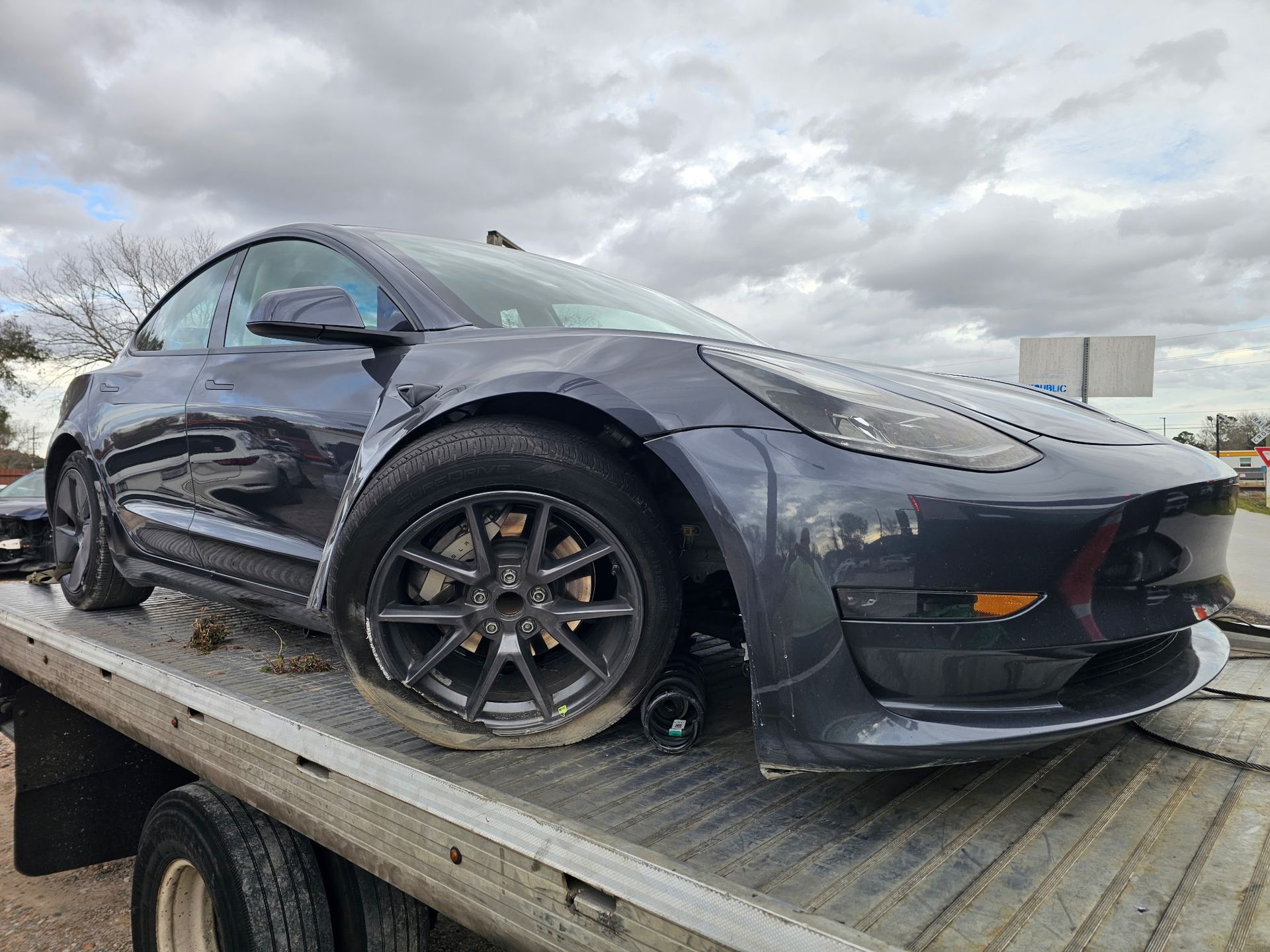 Damaged gray Tesla Model 3 on a tow truck, with front driver's side tire flat and body damage.