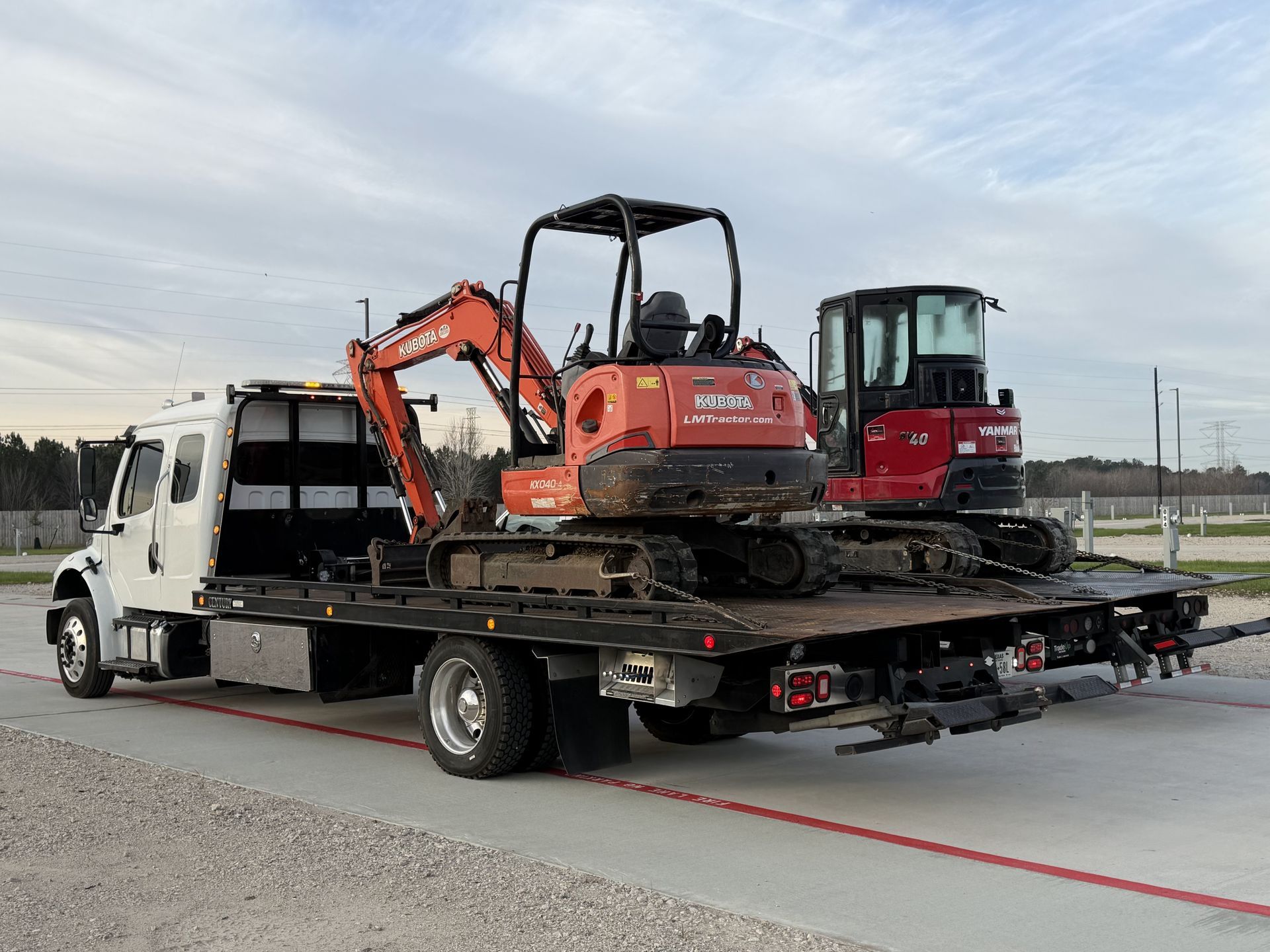 Flatbed truck carrying two excavators on a gray road. Cloudy sky in the background.