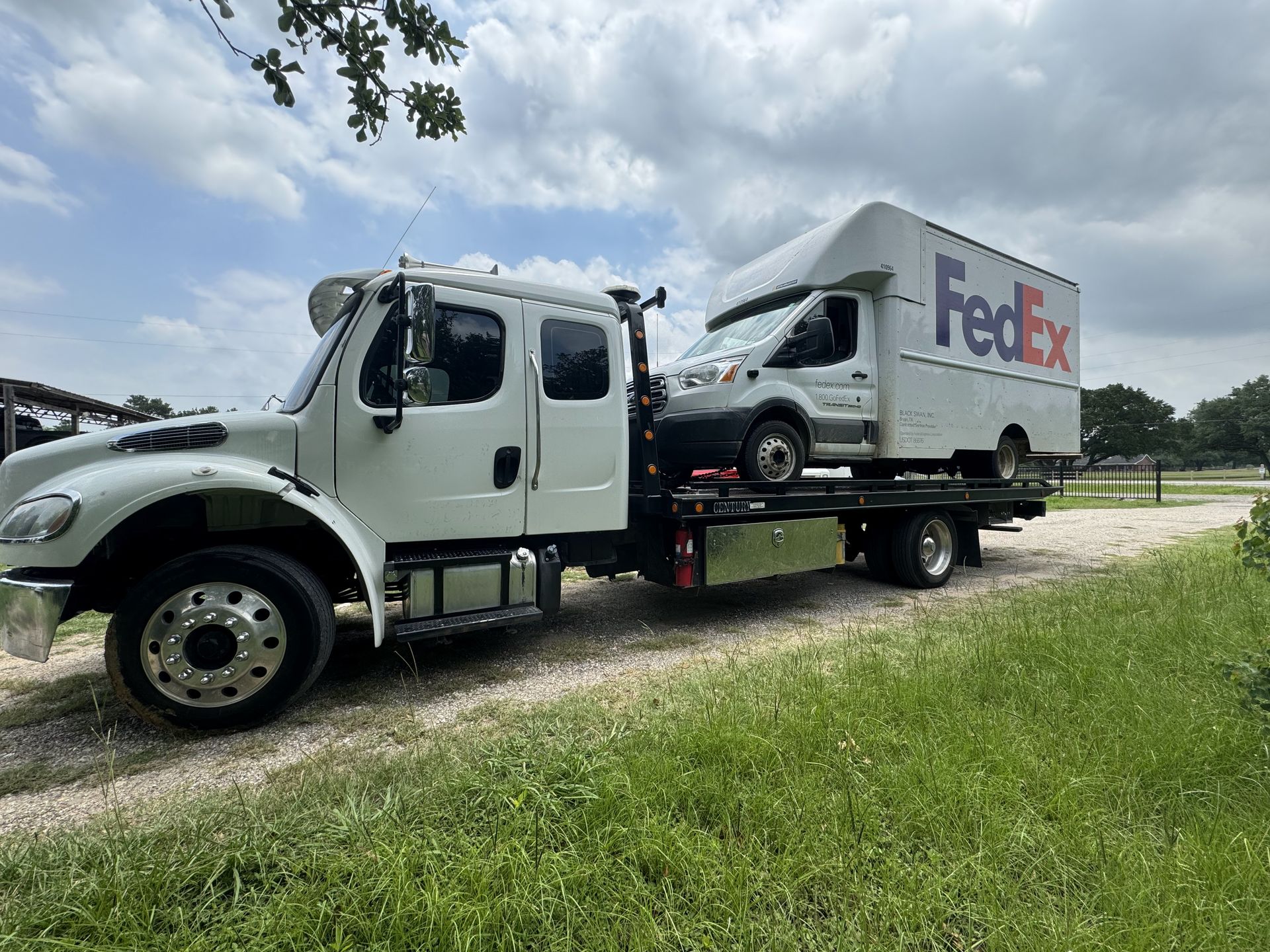 White tow truck carrying a FedEx delivery van on a flatbed, outdoors on grass.