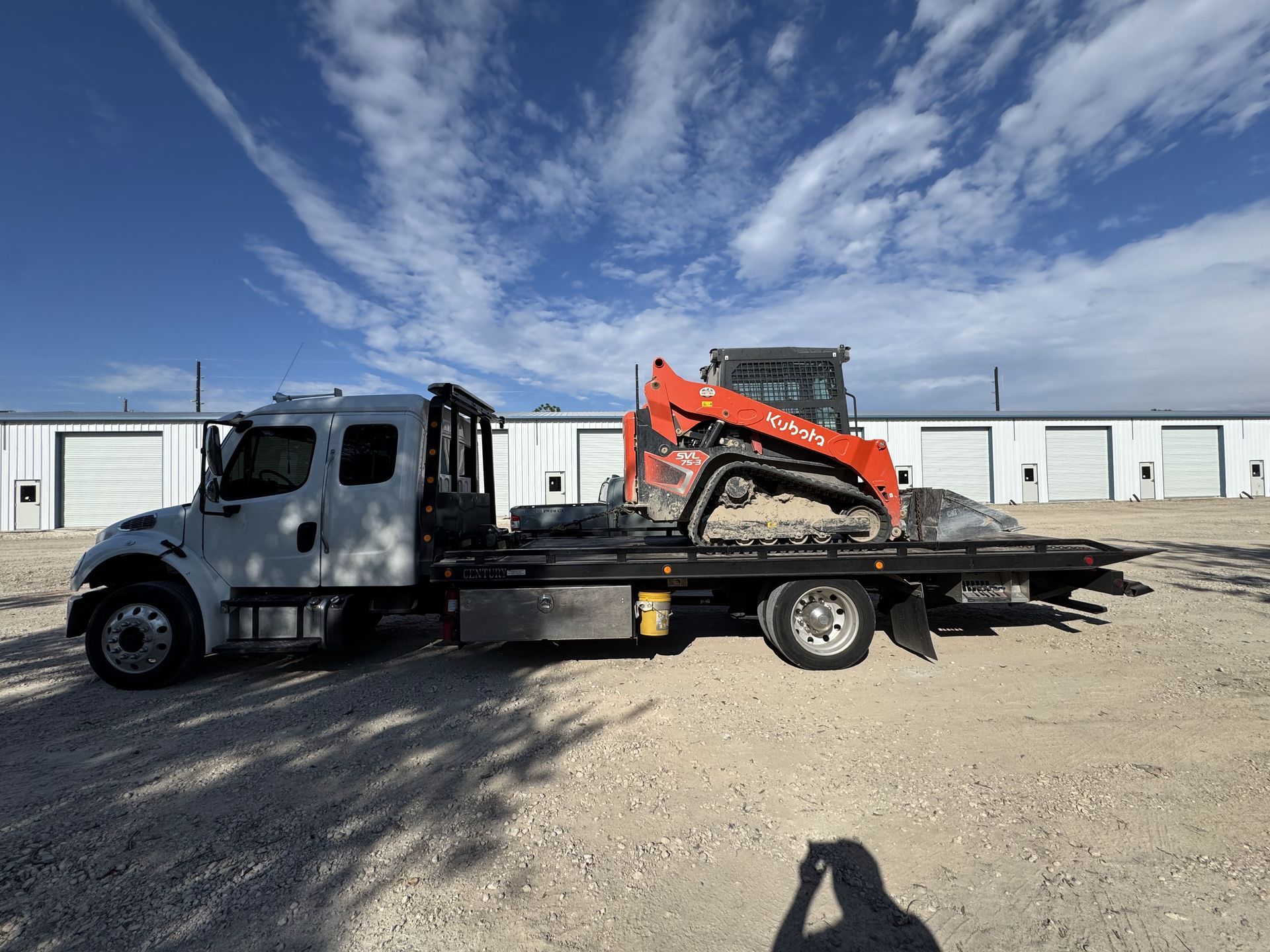 White tow truck carrying an orange Kubota track loader on its flatbed under a partly cloudy sky.